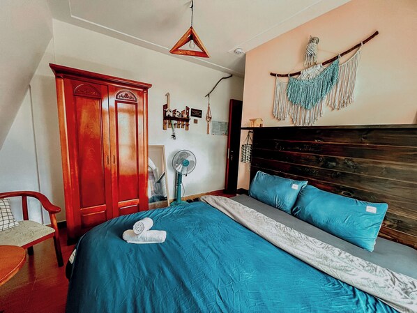 Quarto com cama, guarda-roupa de madeira, peça de macramê na parede e ventoinha no teto.