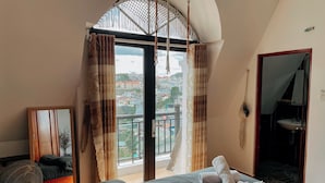 Une chambre avec un lit, un ventilateur, une vue sur la ville depuis un balcon, et une salle de bain visible à travers une porte ouverte.