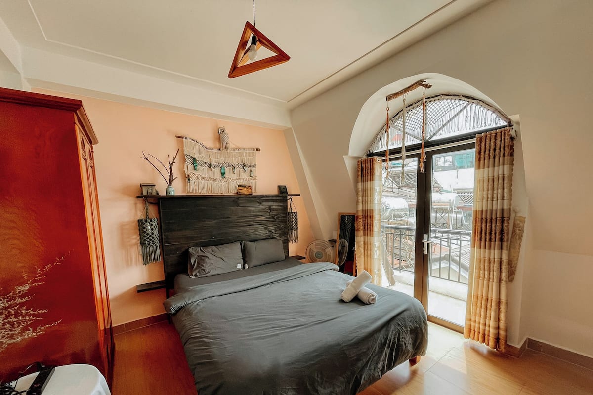 A bedroom with a bed, a wooden headboard, a wall-mounted macrame piece, a fan, and a view of the outside through a window with curtains.