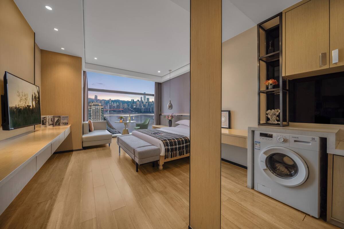 A modern bedroom with a large window, a bed, a washing machine, and a view of the city skyline.