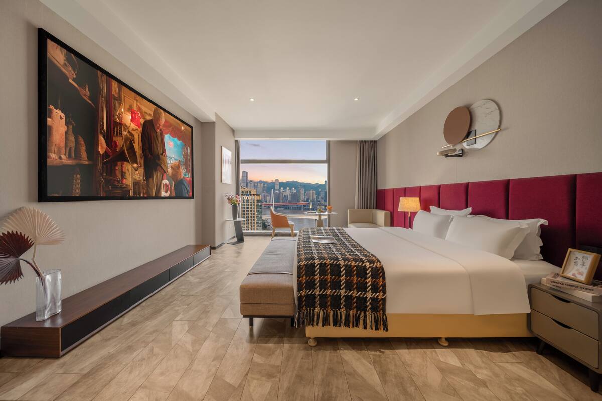 A modern hotel room with a large bed, a city view, a long wooden bench, and a large painting on the wall.