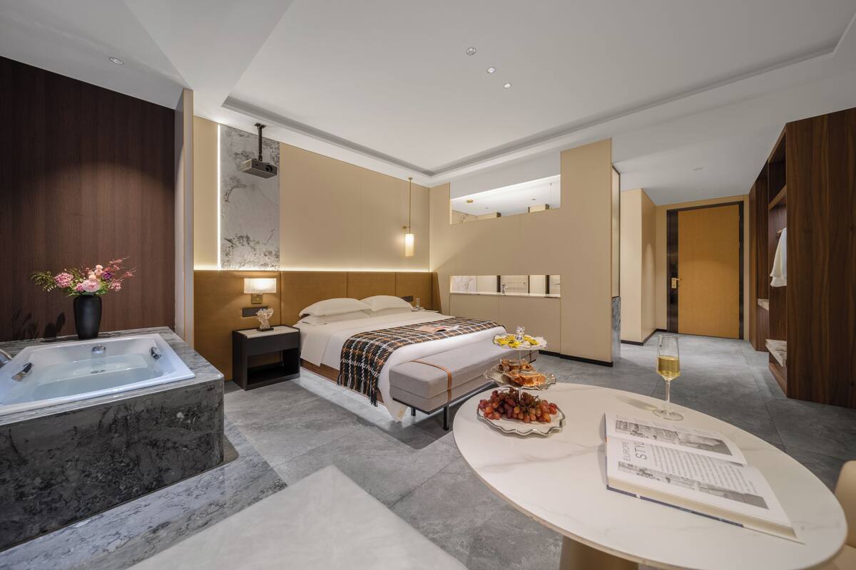 A modern hotel room with a large bed, a separate bathroom with a bathtub, a dining table with a glass of wine, and a small side table with snacks.