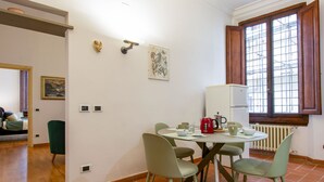 Uma área de refeições com uma mesa redonda, quatro cadeiras, uma chaleira vermelha e uma janela com persianas de madeira.