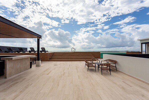A rooftop terrace with a view of the sea, a seating area with chairs, and a bar.