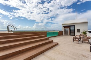 A modern outdoor terrace with wooden steps leading to a seating area, a view of the ocean, and a clear sky.