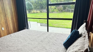 A bedroom with a large window, a bed with a patterned bedspread, and a view of a grassy yard. - Alpaca Farm Cottage (Ongarue)