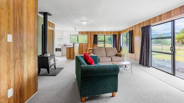 A modern living room with a sofa, armchair, and a fireplace. There is a kitchen area in the background and large windows with curtains. - Alpaca Farm Cottage (Ongarue)