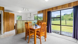 A dining area with a wooden table and chairs, a kitchen with wooden cabinets, and large windows with a view of a green landscape. - Alpaca Farm Cottage (Ongarue)