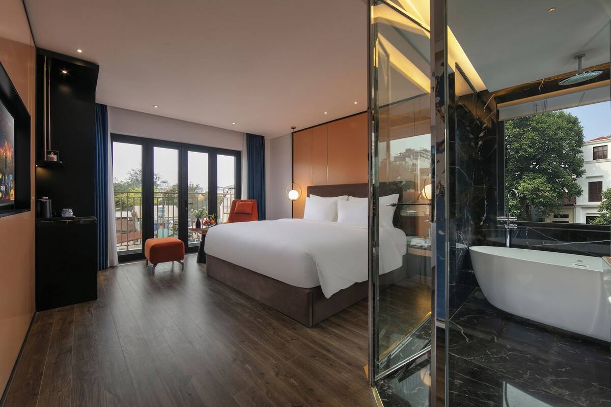 A modern hotel room with a large bed, a glass-enclosed bathroom, and a balcony with a view.