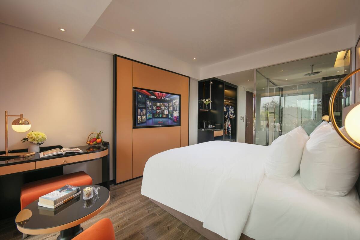 A modern hotel room with a large bed, a desk with a lamp, a small table with a book and a glass, and a wall-mounted TV displaying Netflix content.