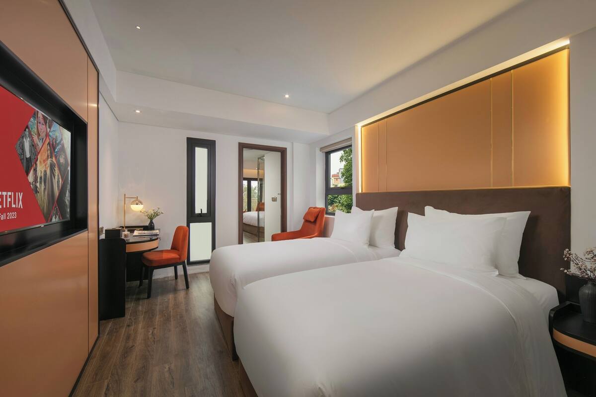 A hotel room with two beds, a desk with a chair, a TV, and a large window.