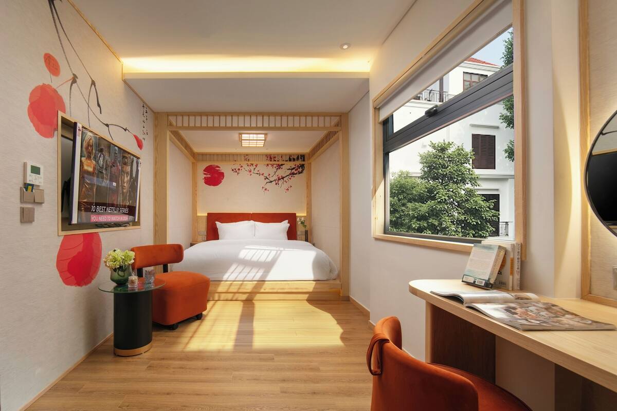 A modern hotel room with a bed, a desk, a chair, and a window with a view of buildings.