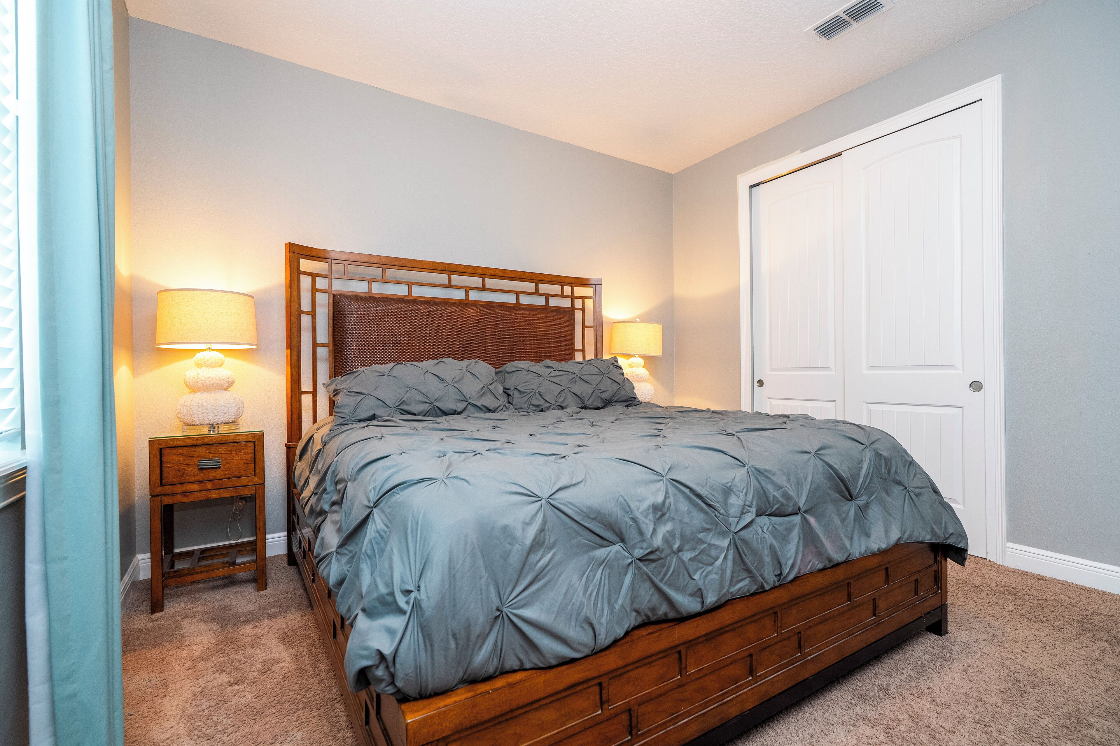 Een slaapkamer met een houten bedframe, een grijze gestoffeerde spreideken, twee bedlampen, een nachtkastje en een witte deur.