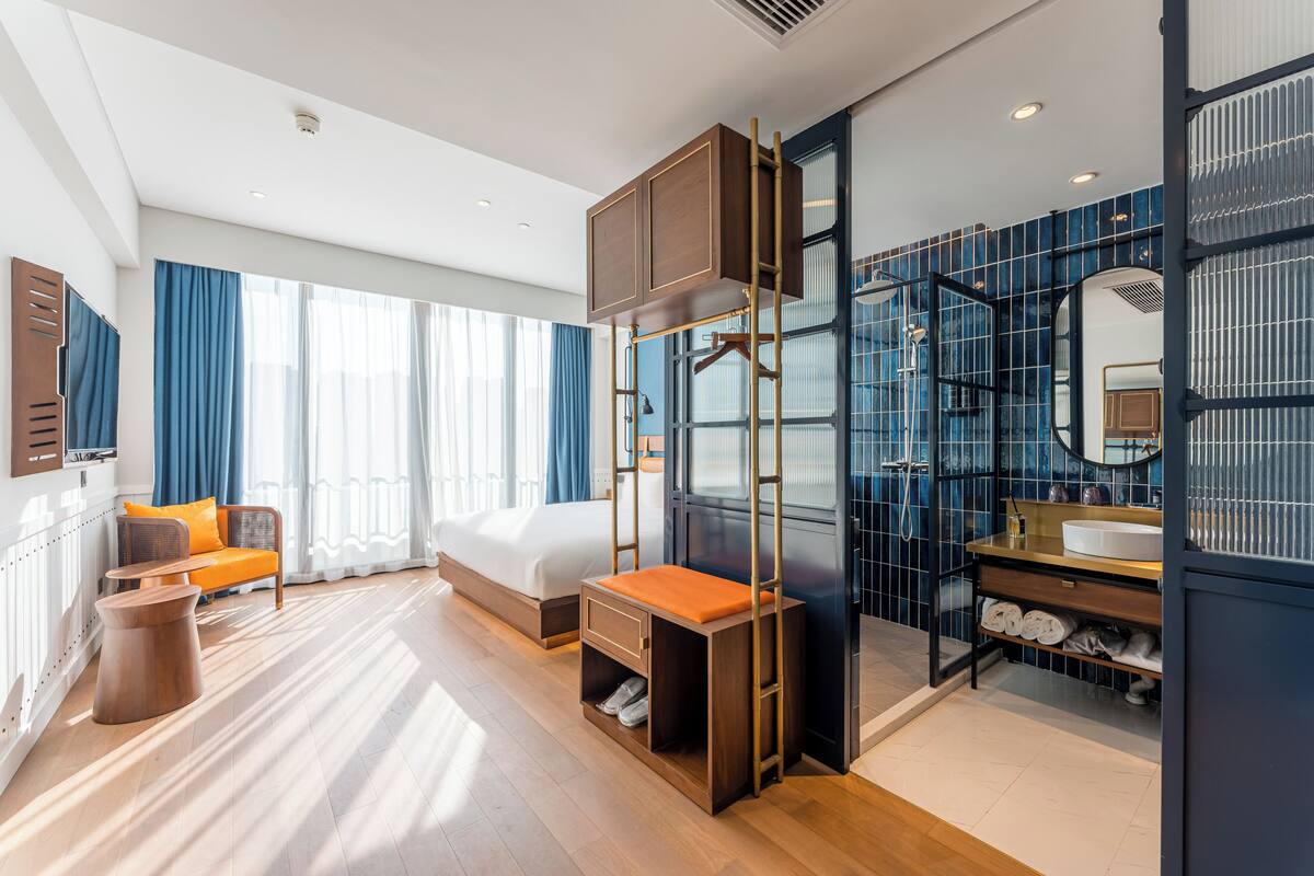 Habitación de hotel moderna con cama, banca de madera, mesita, armario empotrado, baño con espejo y ventana grande con cortinas.