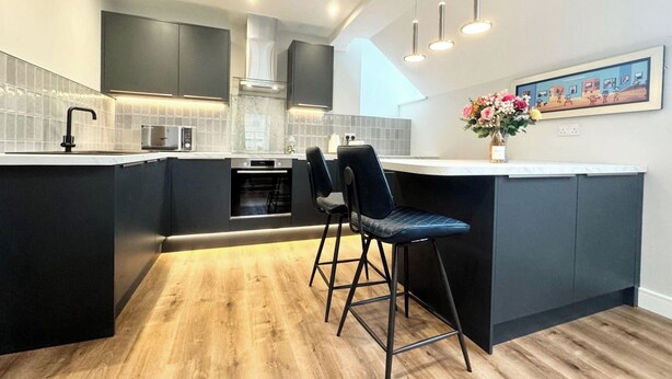 Une cuisine moderne avec des meubles de couleur sombre, un îlot central doté de tabourets, un four encastré et un revêtement mural en carreaux blancs.