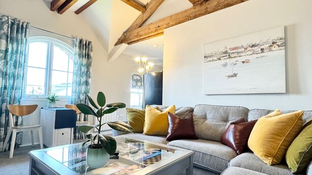 A modern living room with exposed wooden beams, a glass coffee table, a grey sofa with colorful cushions, and a painting of a coastal scene. - Warehouse Apartments (Ulverston)