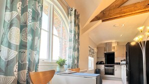 A cozy kitchen with a built-in fridge, a dining area with a wooden chair, and a window with patterned curtains. - Warehouse Apartments (Ulverston)
