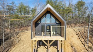 Una baita con veranda in legno e ampie finestre, circondata da alberi e un sentiero sterrato.