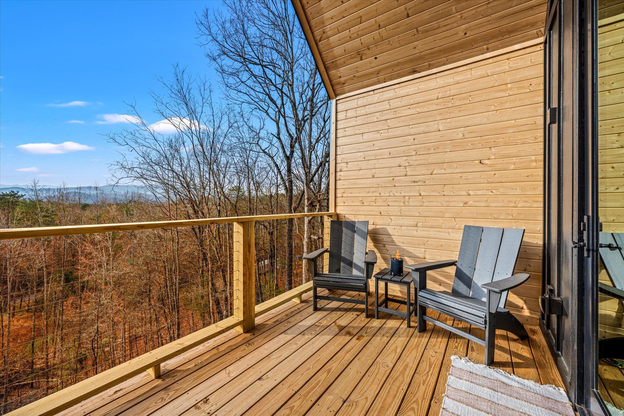 Eine Holzterrasse mit zwei Adirondack-Stühlen und einem kleinen Tisch, mit Blick auf eine bewaldete Gegend.