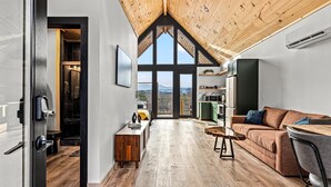 A modern interior with a high ceiling, wooden flooring, a sofa, a dining table, and a view through large windows. - Romantic Luxury Treehouse - Hot Tub, Soaring Views, WiFi, Cable, Mins to PF & GB (Sevierville)