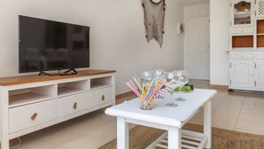 A living room with a flat-screen TV on a wooden TV stand, a white coffee table with a vase of colorful straws and wine glasses, and a white cabinet in the background.