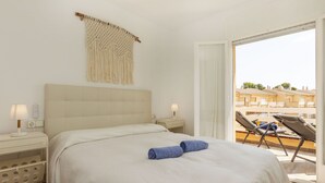 A bedroom with a large bed, two bedside tables with lamps, a macrame wall hanging, and a balcony with chairs.
