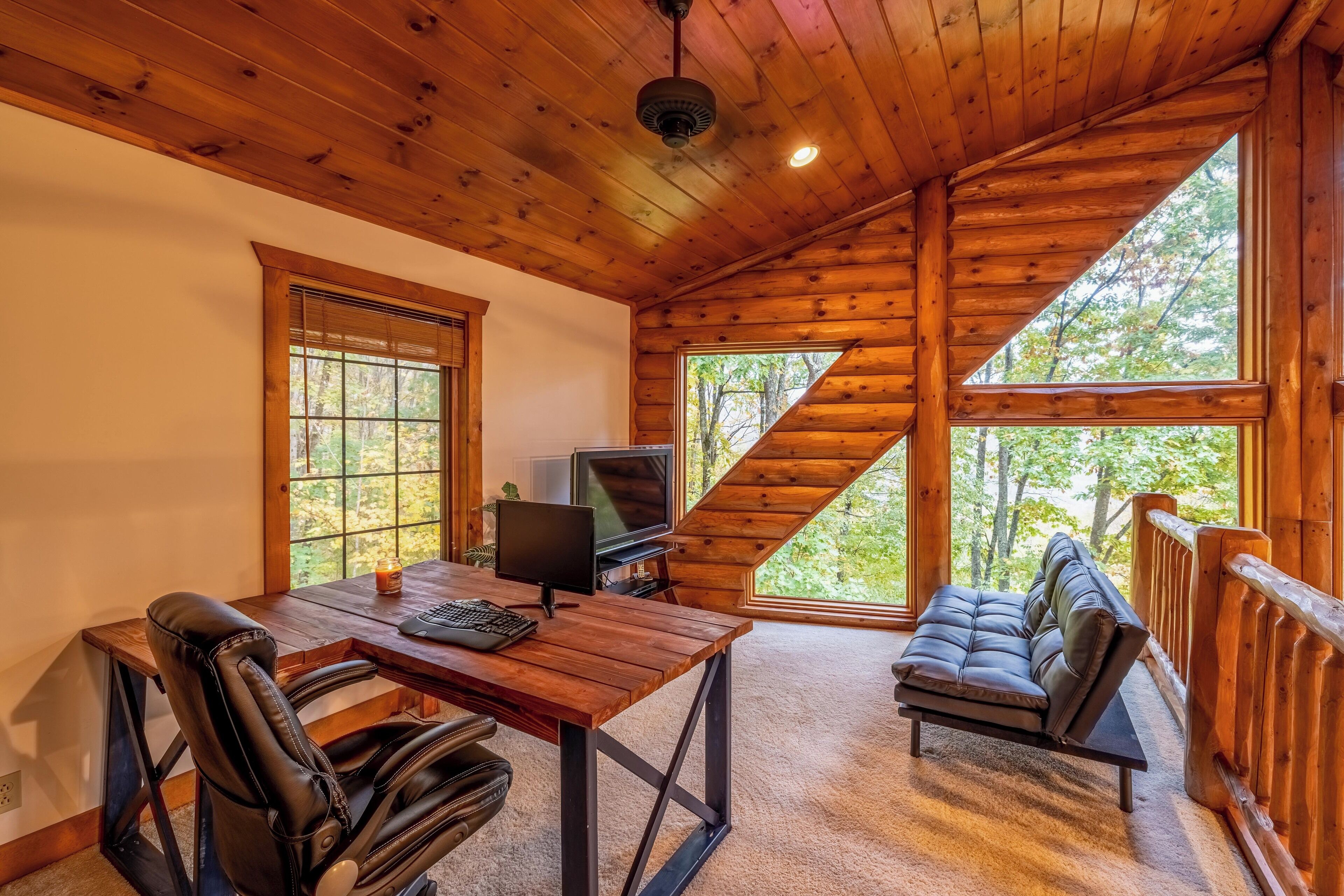 Una cabaña con interior de madera, un escritorio con computadora, un sofá y ventanas amplias con vista a árboles.