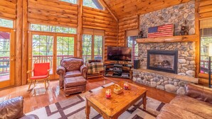 Interior de una cabaña de madera con chimenea de piedra, muebles de cuero y televisor.