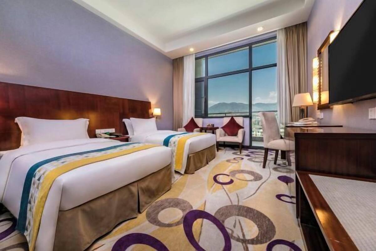A hotel room with two beds, a desk, a chair, a TV, and a large window with a mountain view.