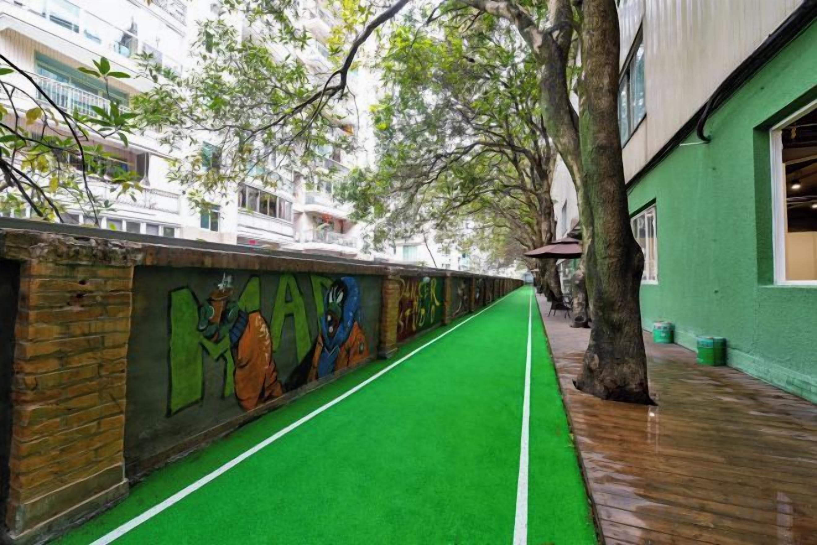 A narrow pathway with green artificial turf, a brick wall with colorful graffiti, and a tree-lined sidewalk.