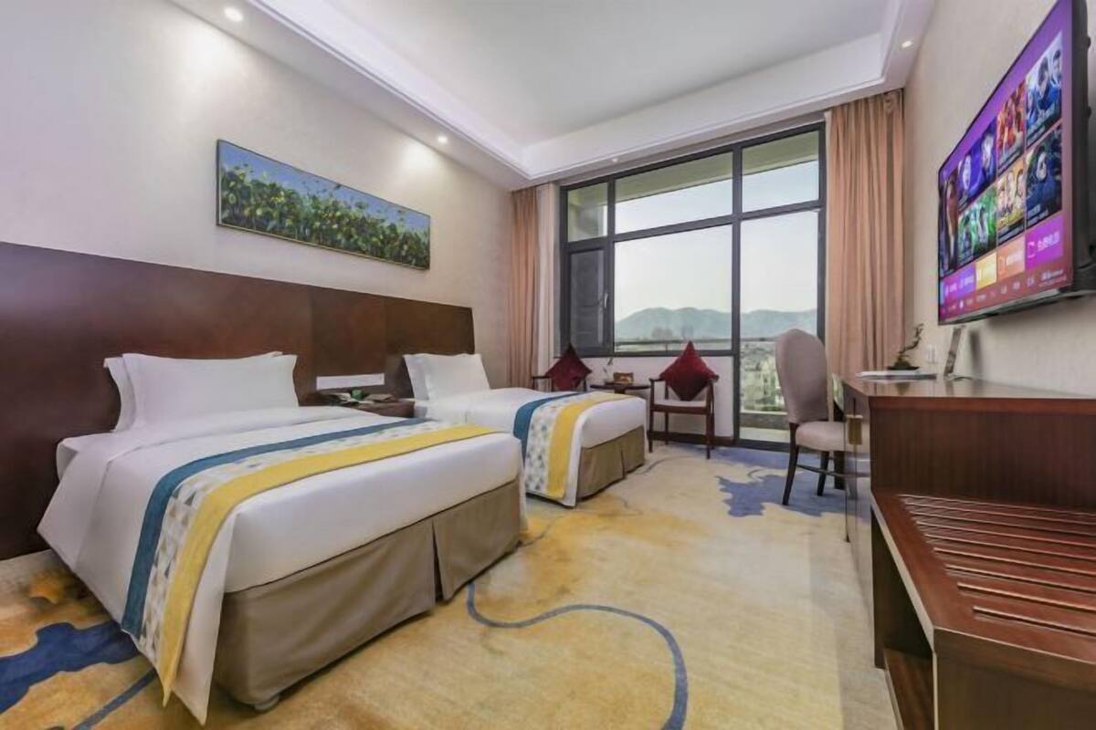 A hotel room with two beds, a desk, a TV, and a large window with a view.