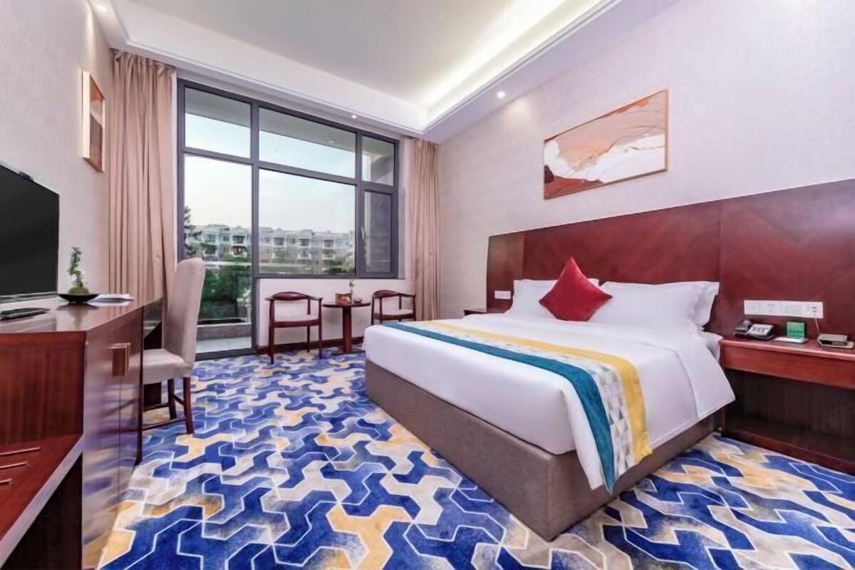 A hotel room with a large bed, a desk with a TV, a small table, and a window with a view of buildings.