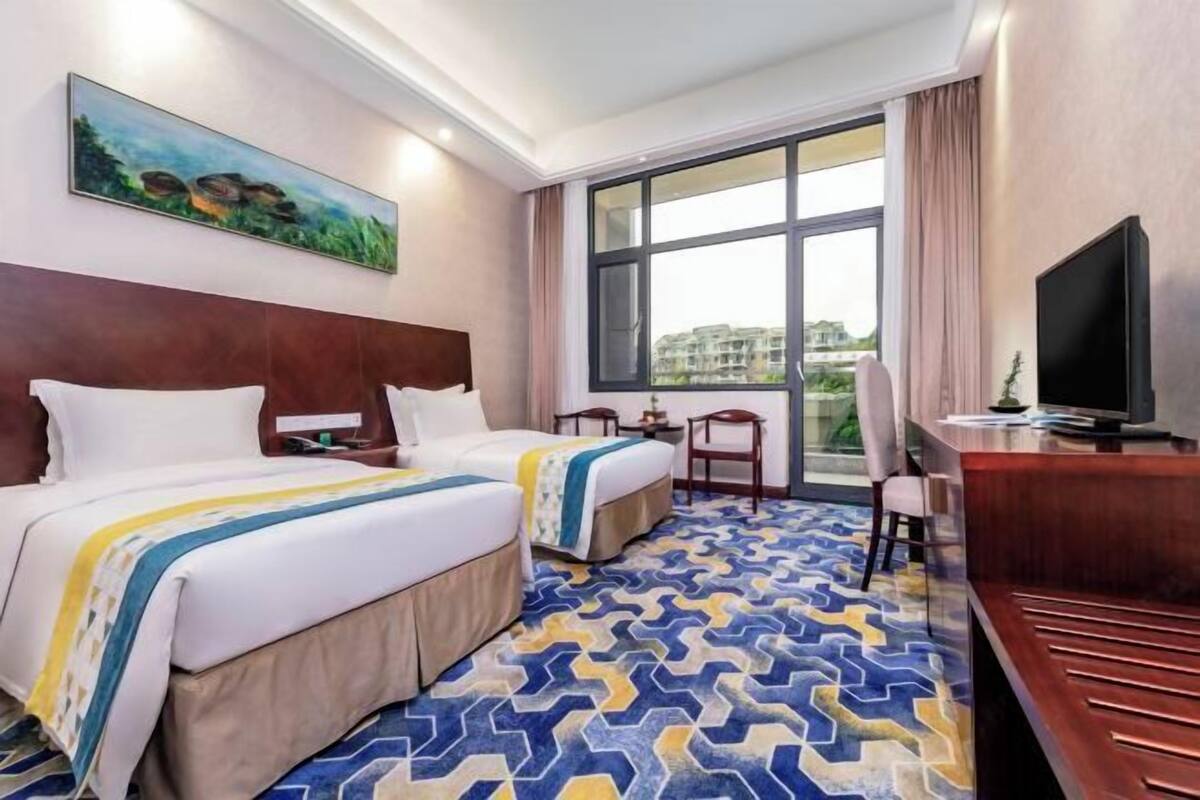 A hotel room with two beds, a desk, a TV, and a large window with a view of buildings.