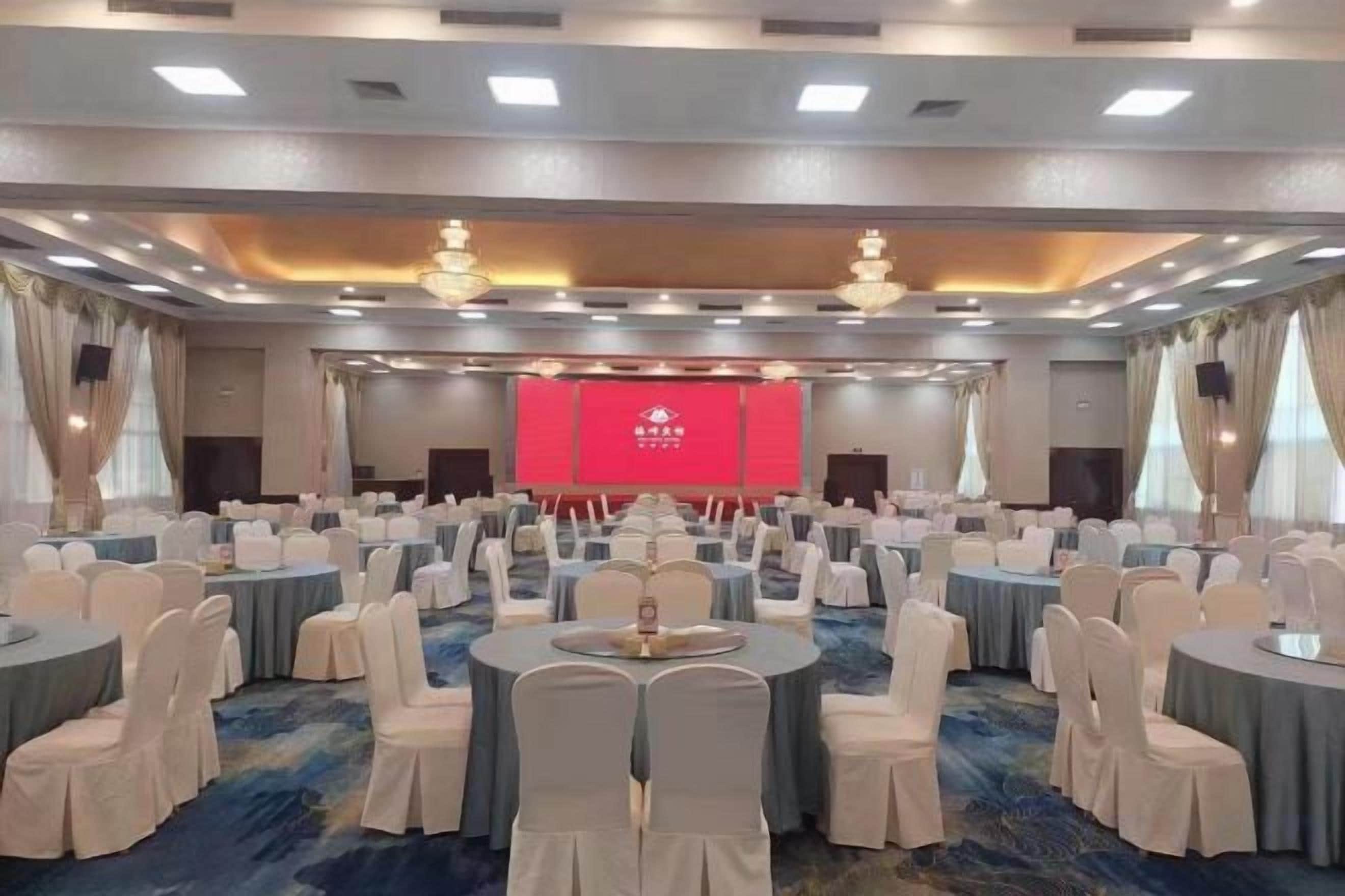 Une salle de conférence avec des tables et des chaises rondes, une scène dotée d’un fond rouge et des lustres.