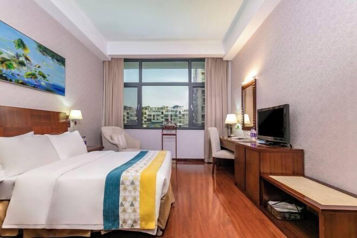 A hotel room with a bed, a desk with a TV, a chair, a window with curtains, and a painting on the wall.