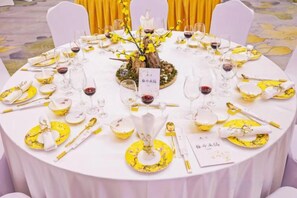 Mesa de jantar redonda posta para uma refeição, com cartões de lugar, copos de vinho e talheres.