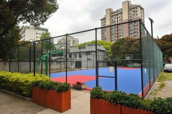 Quadra de basquete ao ar livre com cerca, rodeada por vegetação e edifícios ao fundo.