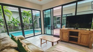 A modern living room with a leather sofa, a wooden coffee table, and a TV on a wooden cabinet. Large glass doors lead to a pool area with lush greenery. - Beautiful 4 Bedroom Private Pool Villa in Laguna Bangtao, Daily Cleaning (Tambon Choeng Thale)
