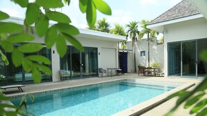 A modern pool area with a clear blue pool, surrounded by glass doors and windows, and furnished with outdoor seating. - Beautiful 4 Bedroom Private Pool Villa in Laguna Bangtao, Daily Cleaning (Tambon Choeng Thale)