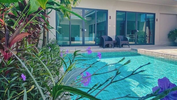 A swimming pool with a glass enclosure, surrounded by lush greenery and purple flowers. - Beautiful 4 Bedroom Private Pool Villa in Laguna Bangtao, Daily Cleaning (Tambon Choeng Thale)