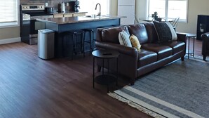 Ein geräumiges Wohnzimmer mit einem braunen Ledersofa, einem kleinen runden Tisch und einer Küchenzeile mit Mikrowelle, Backofen und Kaffeemaschine.
