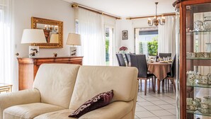Un salon avec un canapé beige, une table d’appoint en bois avec des lampes, un coin repas avec une table et des chaises, et une vitrine contenant des objets décoratifs.