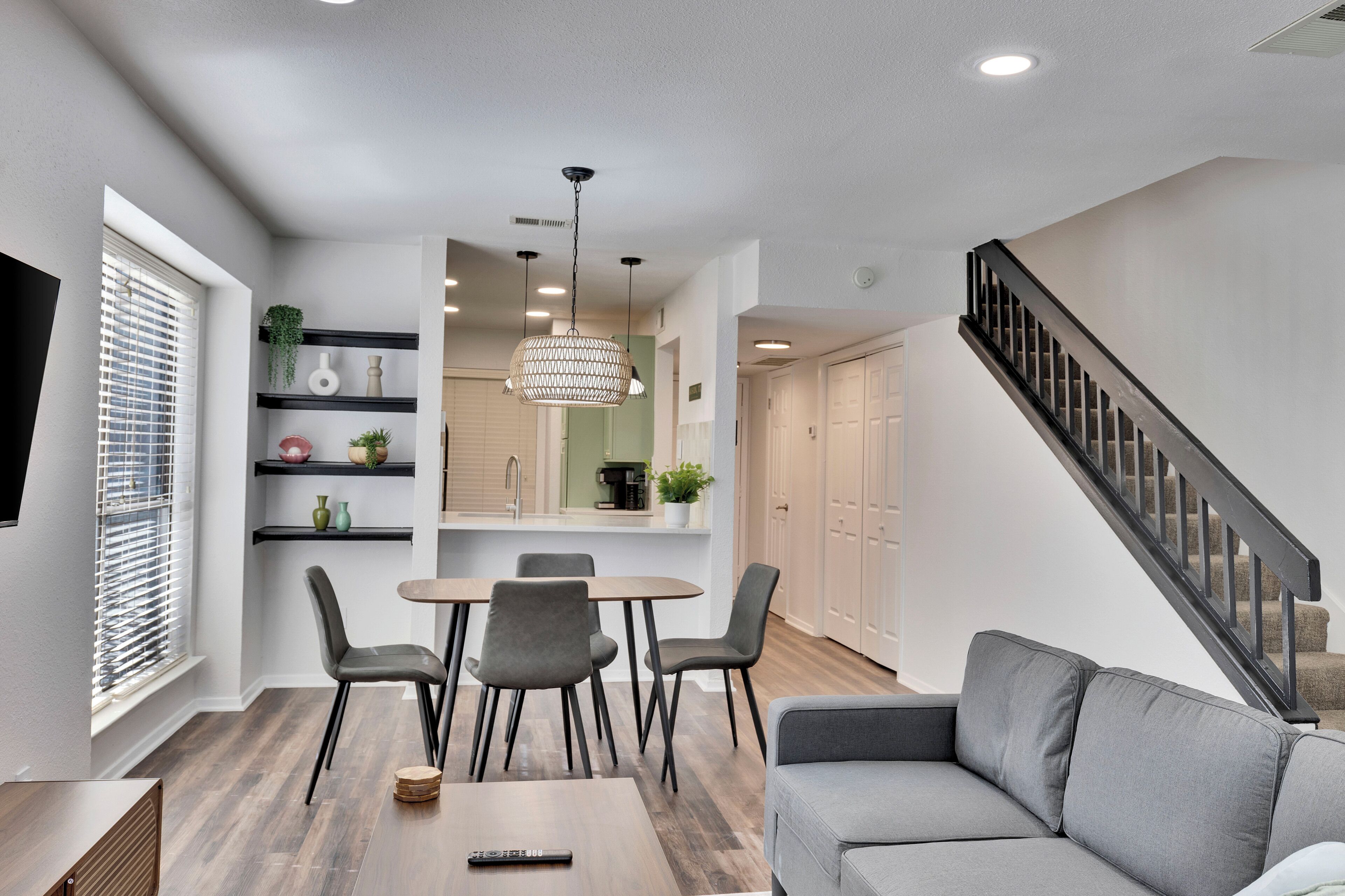Un salon moderne avec un canapé gris, une table à manger avec des chaises, un lustre et un escalier doté d’une rampe noire.
