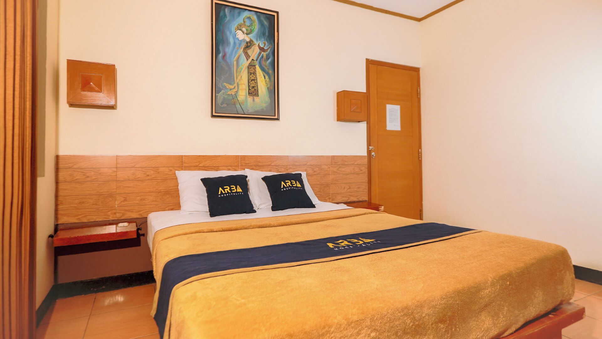 A hotel room with a wooden headboard, a bed with a yellow blanket and two pillows with 'ARBA Hospitality' branding, a painting on the wall, and a wooden door.