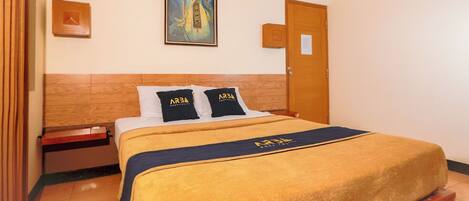A hotel room with a wooden headboard, a bed with a yellow blanket and two pillows with 'ARBA Hospitality' branding, a painting on the wall, and a wooden door.
