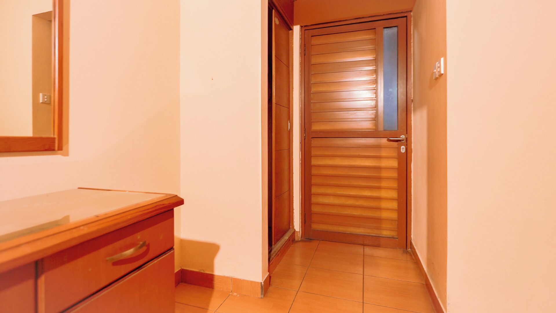 A narrow hallway with a wooden door, a wooden dresser with drawers, and a mirror on the wall.