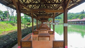 A covered wooden pavilion with rows of benches facing a body of water, surrounded by greenery and a distant building. - Lembah Gunung Kujang by ARBA (Cijambe)