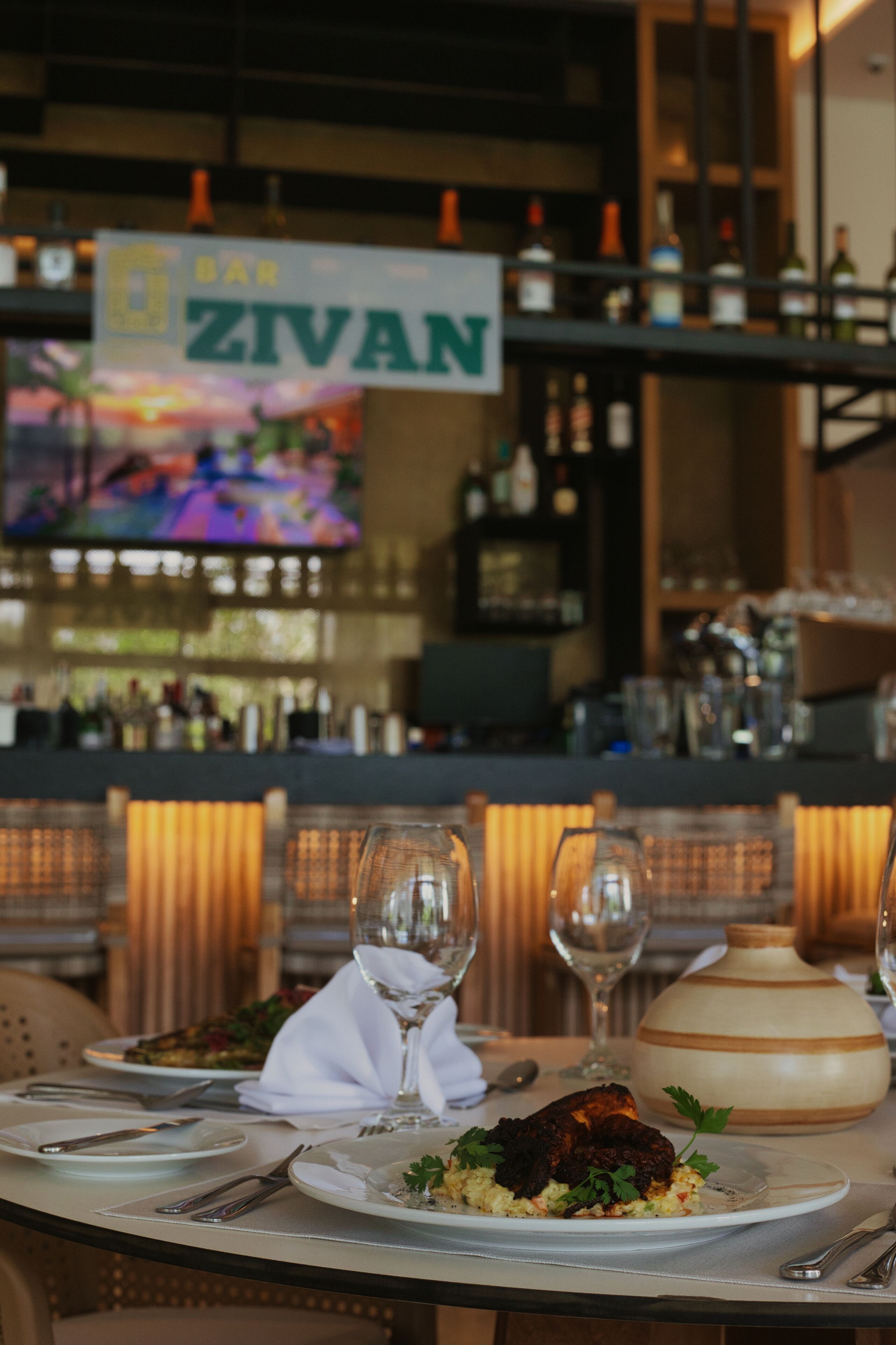 A restaurant setting with a bar area, a television screen, and a table set with plates, glasses, and a vase.