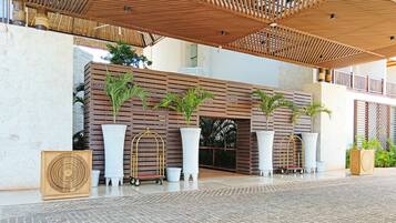 A modern building entrance with a wooden slatted roof, large potted plants, and a white wall with horizontal lines.