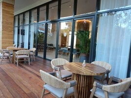 Un amplio espacio al aire libre con mesas de madera y sillas de mimbre, ventanas grandes y vistas del paisaje circundante.